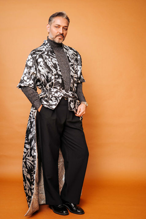Gianni Jovanovic trägt einen auffälligen schwarz-weißen Mantel im Kimono-Stil. Darunter hat er einen glitzernden Pulli und eine schwarze Hose an. Seite Haltung ist aufrecht und souverän. Eine Hand hat er lässig in die Hosentasche gesteckt.