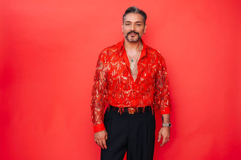 Gianni Jovanovic trägt eine auffällige, glitzernde, rote Bluse. Sie ist tief aufgeknöpft, so dass die Brusthaare und mehrere Goldketten zu sehen sind. Gianni hat grau-melierte Haare und einen Vollbart.