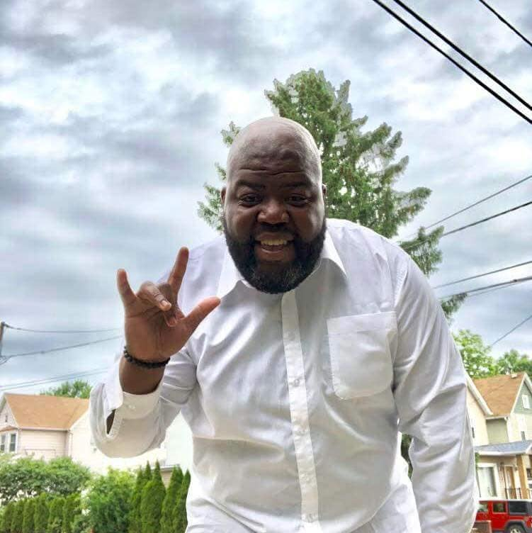 A Black man in a white shirt signs I love you in American Sign Language. He is smiling and standing outside on a cloudy day.