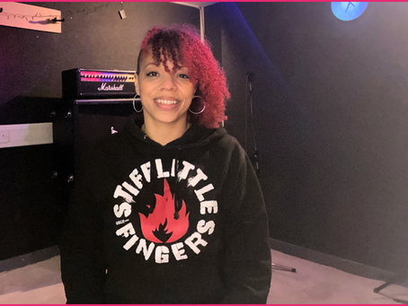 Carrie Griffiths in music studio, smiling at the camera. Carrie has dyed red hair, shaved on the right hand side, wearing a Stiff Little Fingers hoodie.