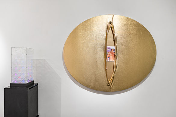 Installation view of Kinky Room, featuring a round golden sculpture with a vertical slit revealing an image inside, flanked by a light-based work on a black pedestal. Works by Katinka Huang, Julio Le Parc, Syd Krochmalny, and Natacha Voliakovsky.