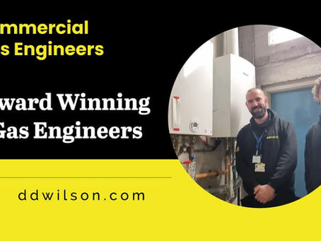 Two gas engineers in hoodies stand smiling beside a boiler in a utility room. Text: "Commercial Gas Engineers, Award Winning Gas Engineers, ddwilson.com".