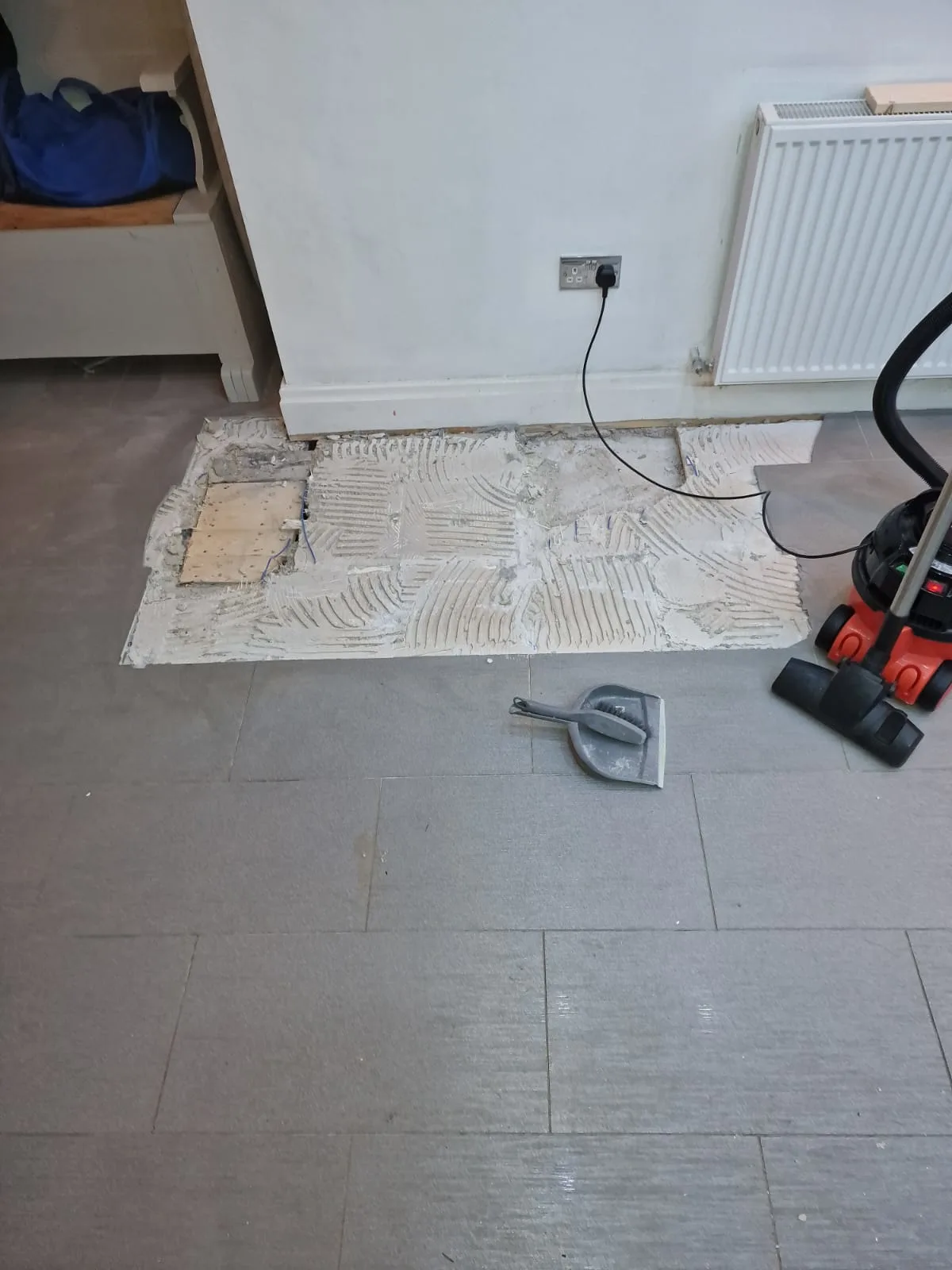 The person taking the picture has stepped back from where the tiles have been lifted up, showing where the tiles are in the kitchen by a radiator. A hoover and brush and shovel are in view.