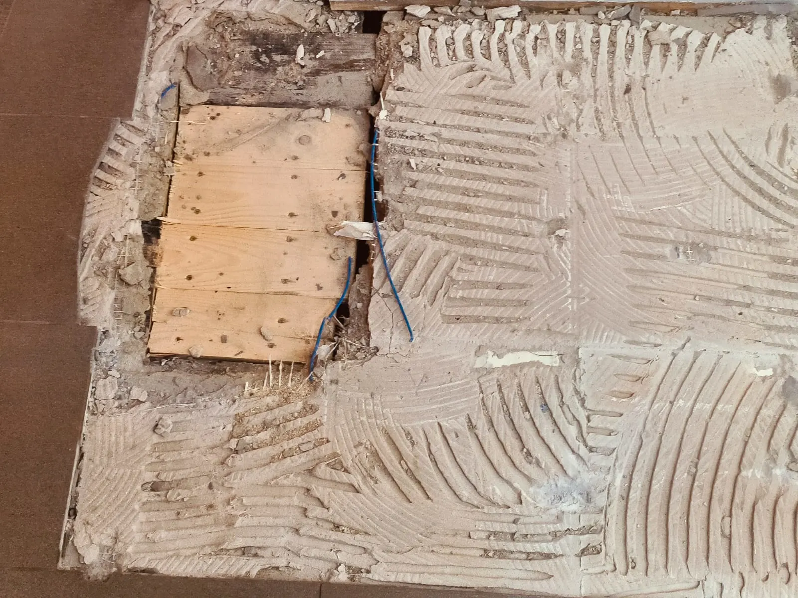 Tiled floor with some tiles removed, exposing the underfloor ring. A piece of wood laid on top of the hole.