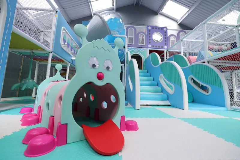 Large play area with pastel colours. A large caterpillar with a clear dome in it's mouth and large red tongue poking out the caterpillar has red wellington boots.