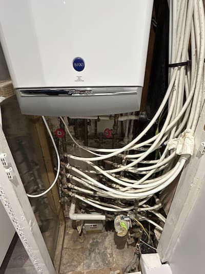 White boiler with multiple white pipes in a small utility space. Visible brands and labels: Baxi, NEST. Worn floor, un-organised pipes. Before works carried out image