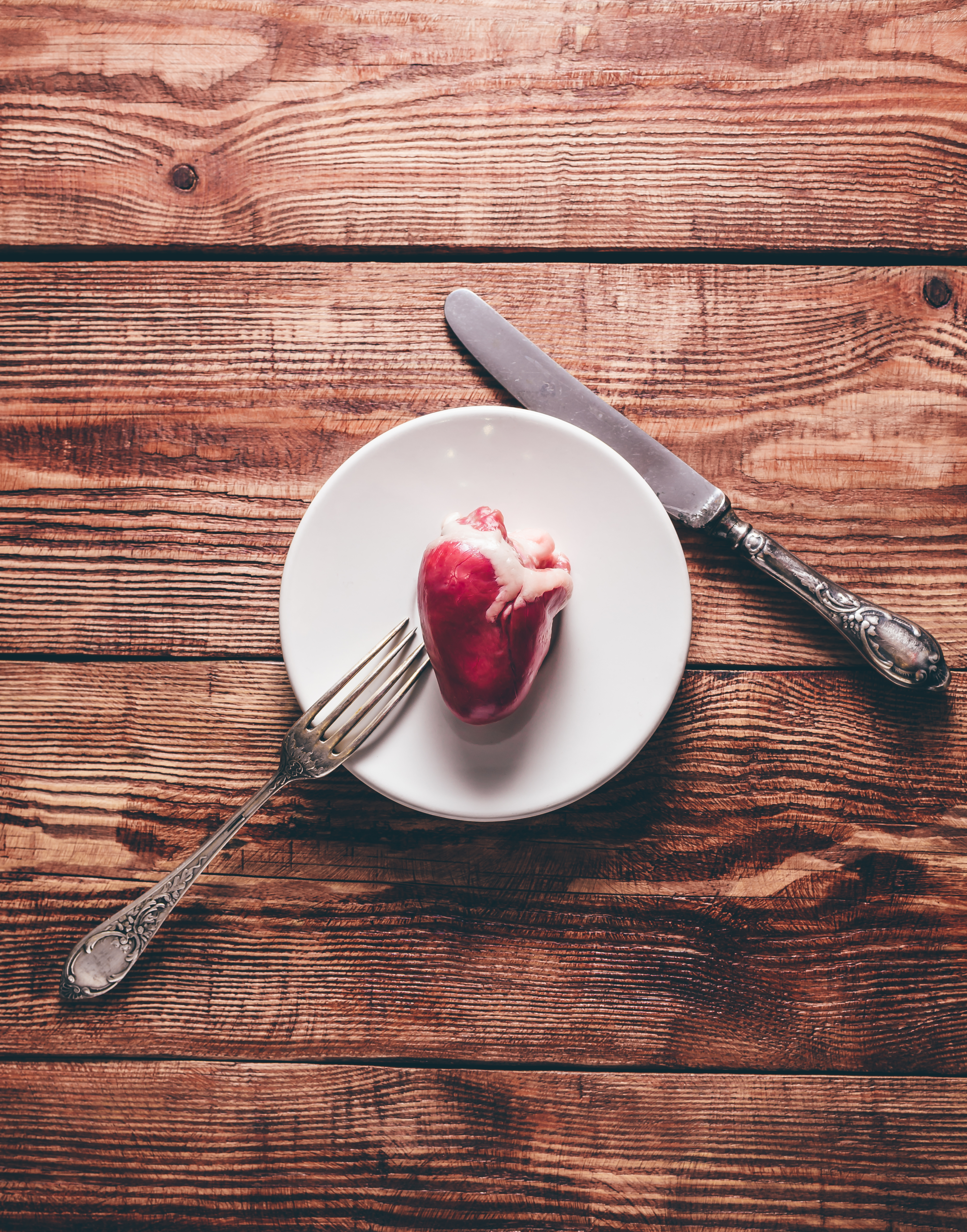 Piccolo cuore rosso crudo su un piatto bianco, con forchetta e coltello d’argento, appoggiato su un tavolo di legno rustico.