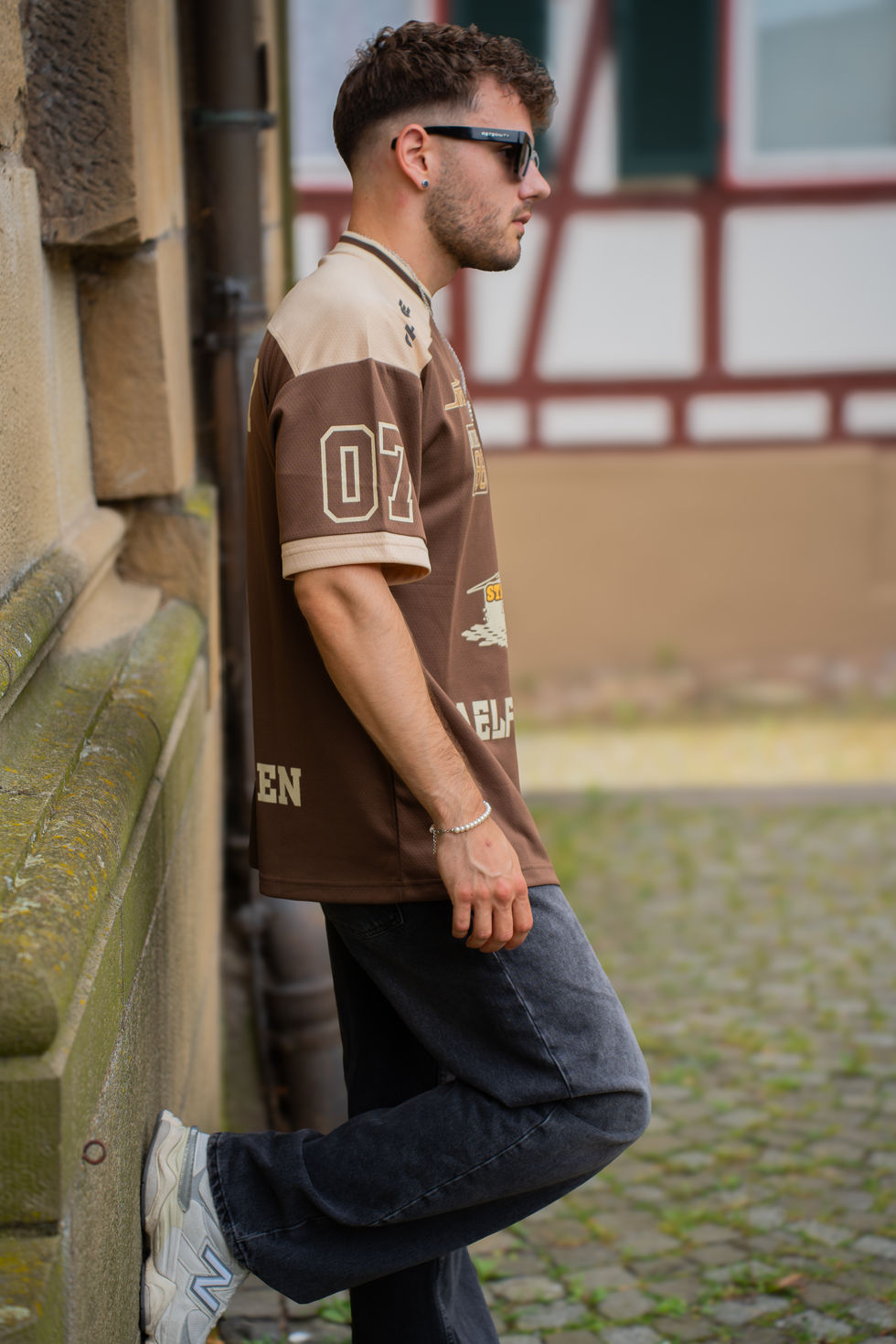Mann lehnt seitlich an Mauer, trägt braunes Shirt und Sonnenbrille.