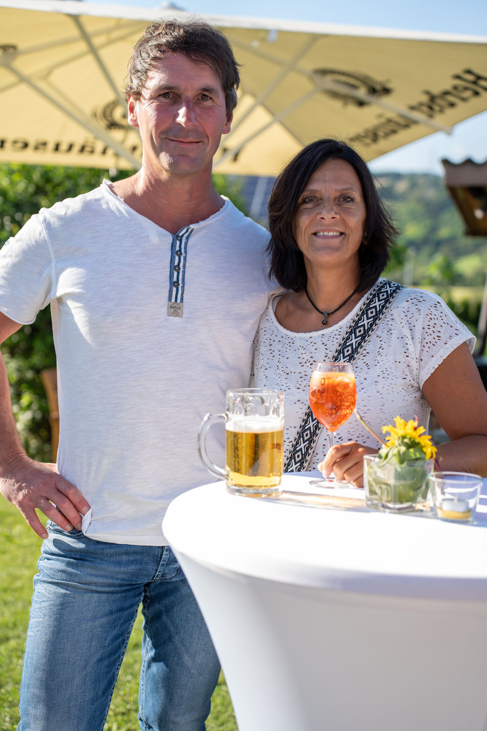 Paar steht mit Getränken an einem Stehtisch und lächelt in die Kamera.