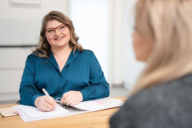Eine freundliche Buchhalterin mit Brille in einer petrolfarbenen Bluse sitzt an einem Schreibtisch. Sie lächelt und hält einen Stift in der Hand, während sie in einem geöffneten Ordner arbeitet. Im Vordergrund ist eine unscharfe Person erkennbar, die ihr gegenüber sitzt. Die Szene vermittelt eine professionelle und einladende Arbeitsatmosphäre.