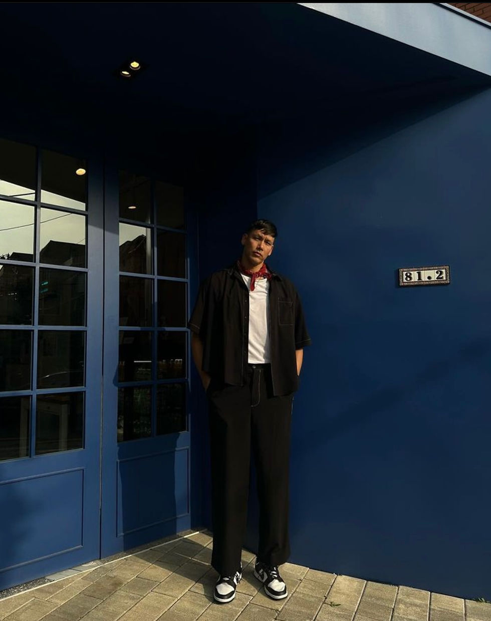 A stylist photo of Eric Sandrin in modern streetwear, wearing a black outfit with sneakers, standing against a vibrant blue wall.