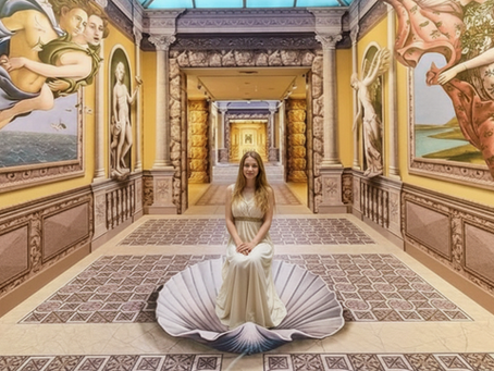 A blonde woman wearing a long white dress sits posed on a large 3D seashell prop in the center of an illusion art museum hallway, recreating the scene from 'The Birth of Venus'.