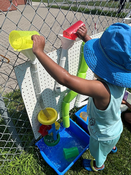 Toddler in blue hat pouring water from cups into play table.