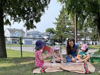 Woman reading to three young children on a picnic blanket outdoors.