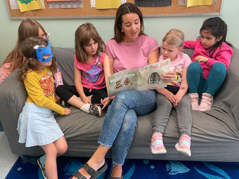 Smiling woman reads storybook to five young children on a couch.