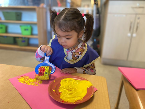 Young child using a roller to paint yellow art on pink paper.
