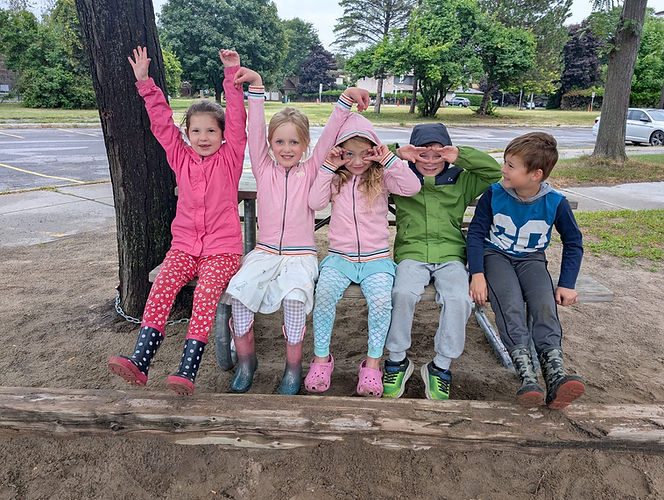 Five playful children making funny faces on a wooden beam outdoors.