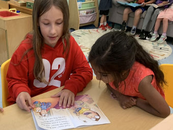 Two girls in a classroom reading a colorful picture book together.