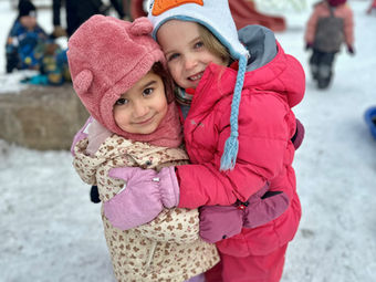 Two young girls hugging, smiling in winter wear, snowy playground background.