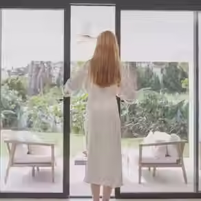 Woman in white robe opening black-framed sliding patio doors to a lush backyard garden.