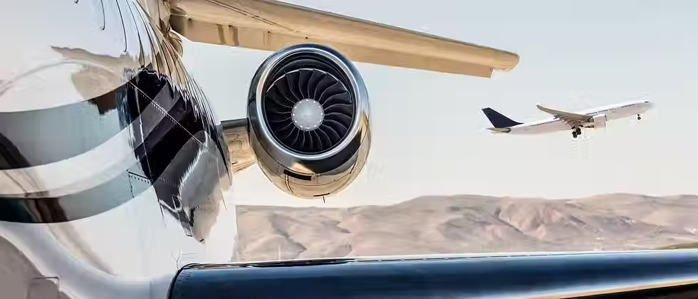 A close-up of a jet engine on a parked aircraft with another plane taking off in the background against a mountainous landscape.