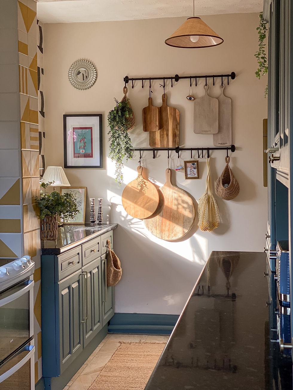 Cozy kitchen with cutting boards on wall, green cabinets, plants, and sunlight creating warm tones. Textured tiles and woven baskets add charm.