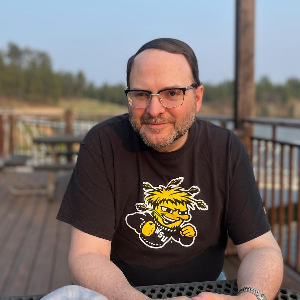 Man with glasses and black t-shirt sitting outdoors.