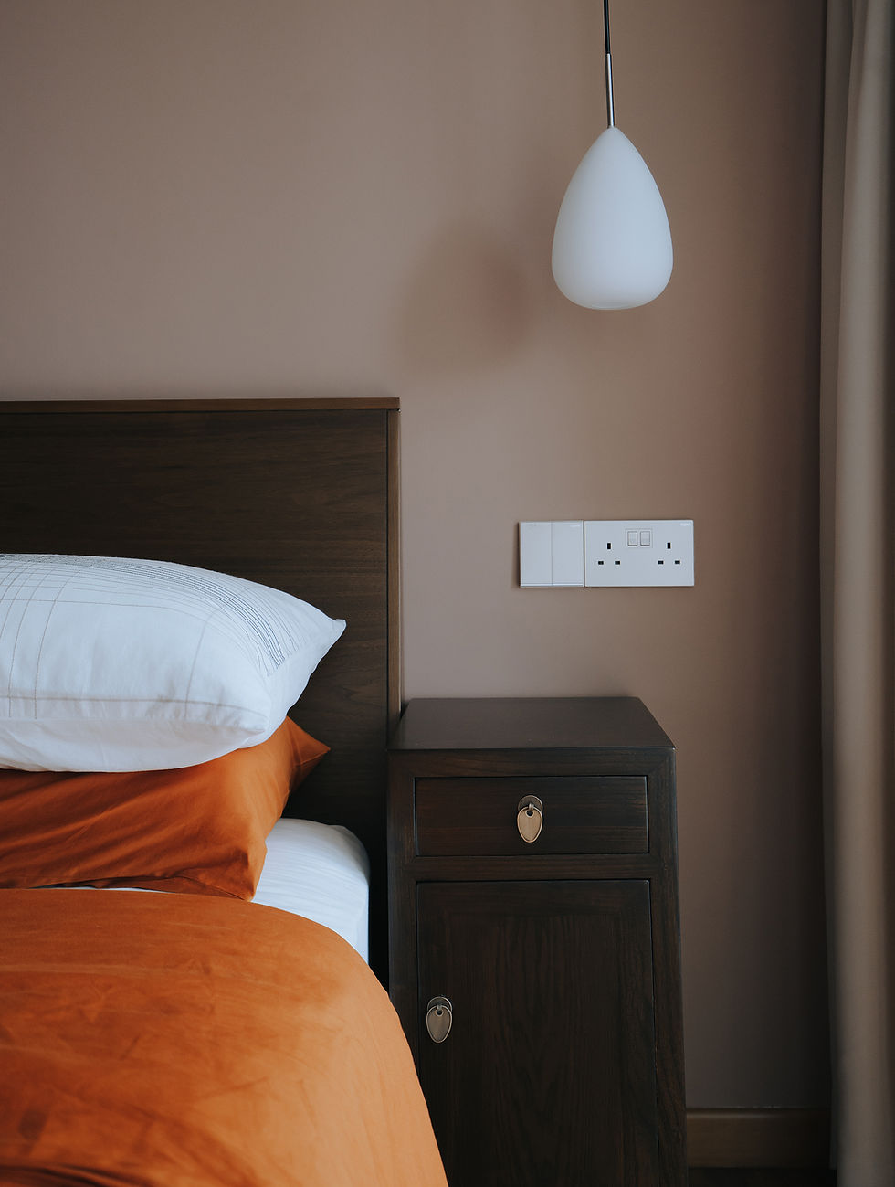 Bedroom scene with a wooden headboard, orange bedding, white pillow, and bedside table. A white pendant light hangs above. Calm ambiance.