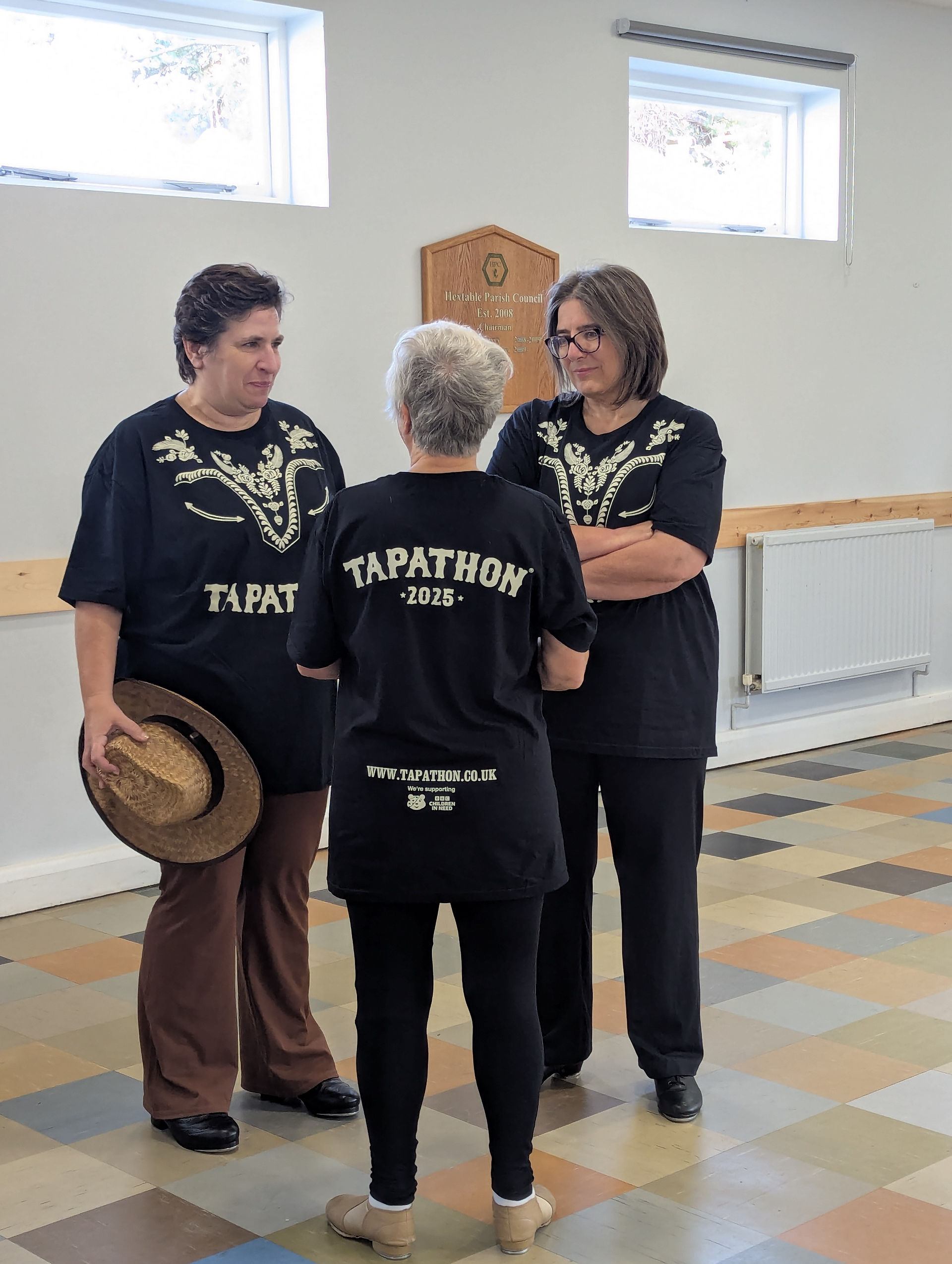 Three Tapathon dancers chatting before the routine at Hextable Village Hall, wearing matching Tapathon 2025 T-shirts and holding cowboy hats.