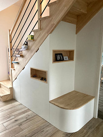 Aménagement sous escalier en bois et médium à peindre sur mesure. Optimisation d'un espace sous escalier avec des rangements sur mesure, penderie, rangement à chaussure et étagères . Cet aménagement fonctionnel intègre des niches décoratives et une assise, offrant un design pratique et esthétique. Parfait pour maximiser l'espace dans une maison moderne tout en apportant une touche chaleureuse avec des finitions en bois.