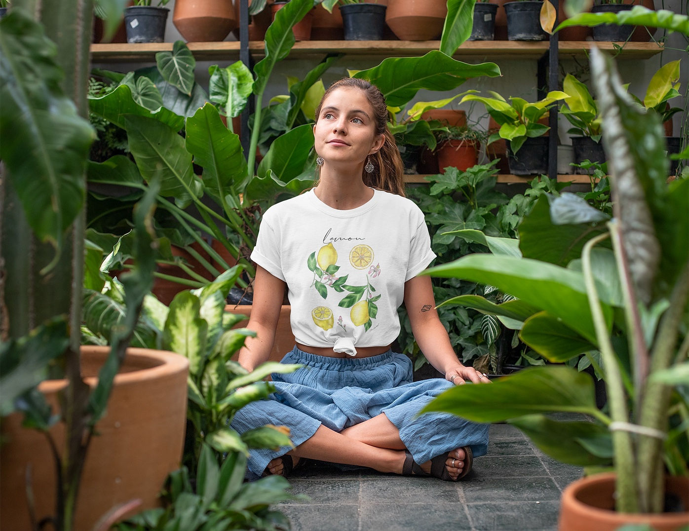 A woman in a white lemon-print t-shirt meditates amidst lush green plants.
