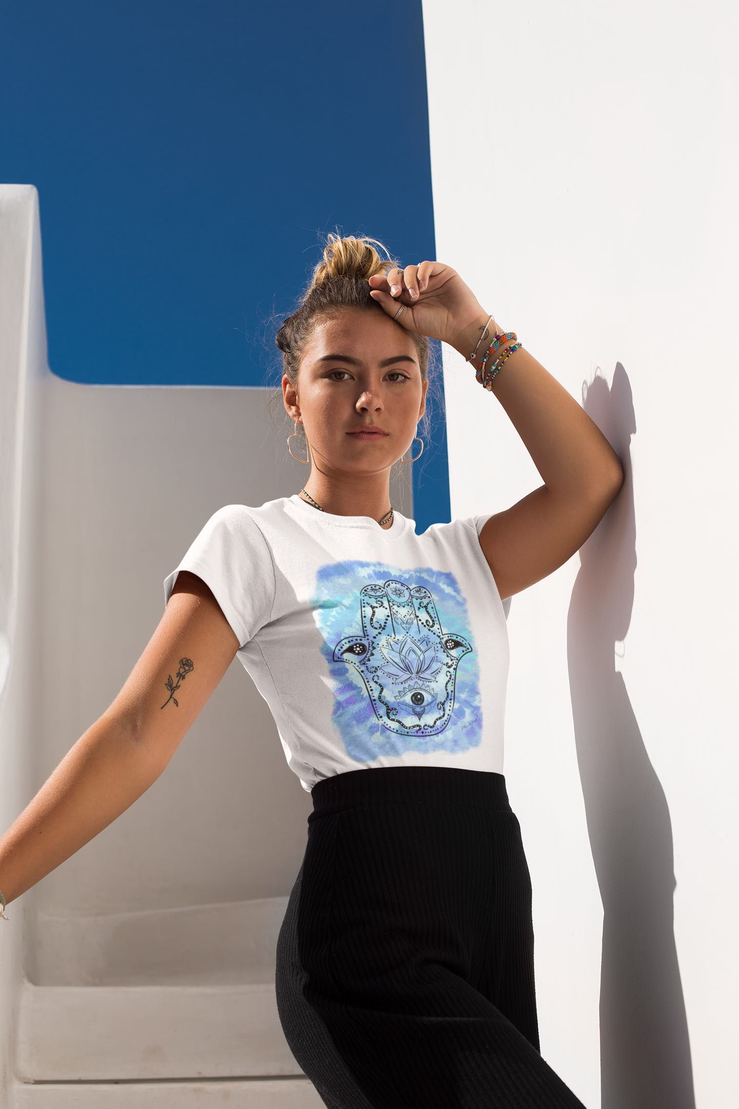 Woman in a Tie Dye Hamsa Hand T-shirt, leaning against a blue-white wall.