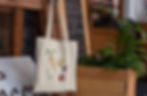 A cream tote bag with carrot, potato, onion, and beet illustrations hangs outdoors.