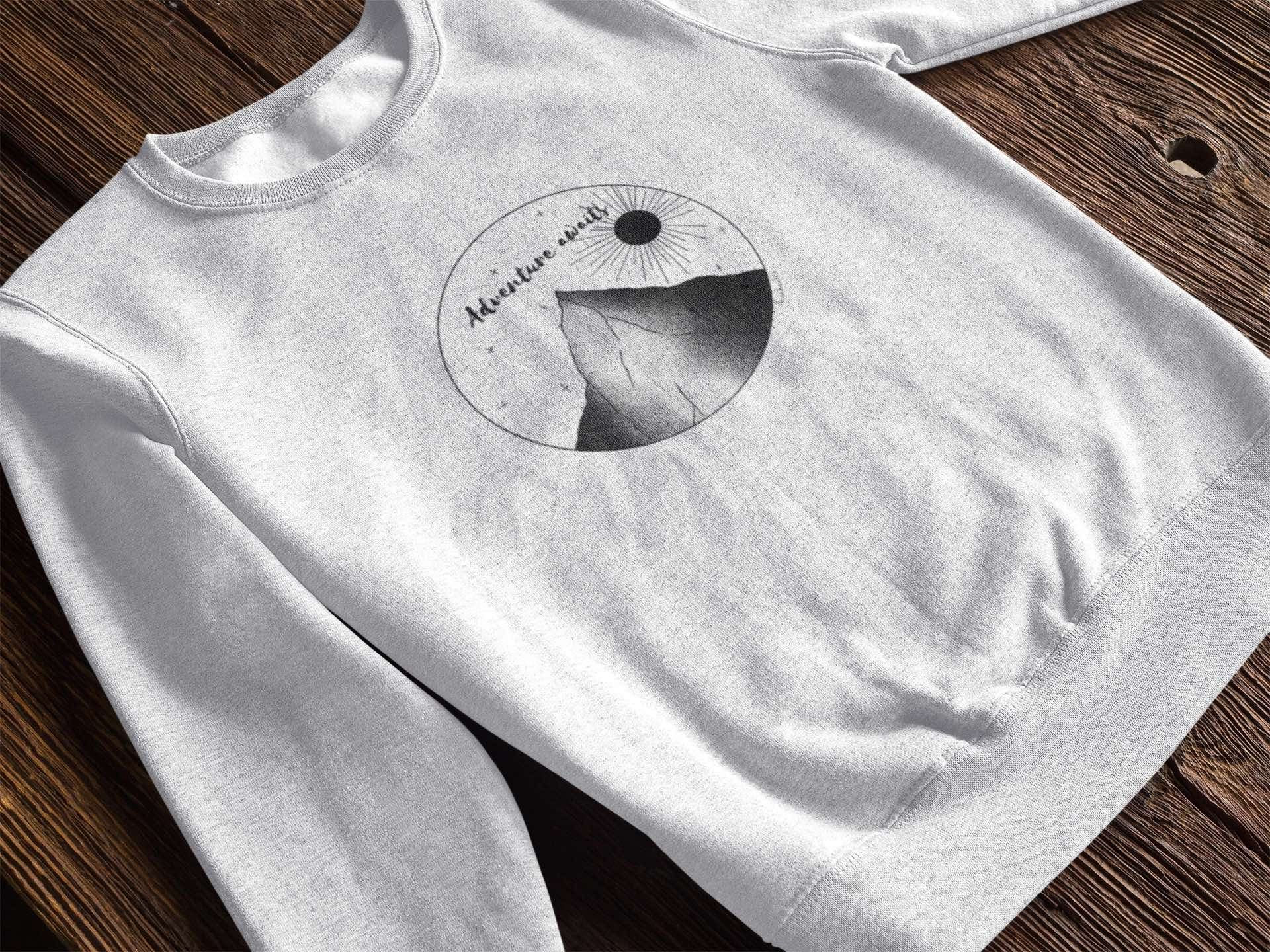 Grey sweatshirt with "Adventure Awaits" text and a greyscale mountain and sun graphic.