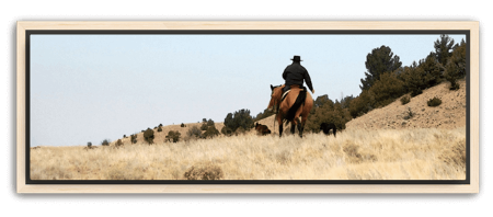 Cowboy on horseback riding through dry grass hills with scattered trees under a clear sky, set in a natural wood frame