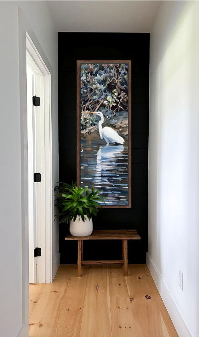 Thumbnail: The same image hanging on a dark wall in a narrow hallway with a door and plant-filled vase on a bench below.