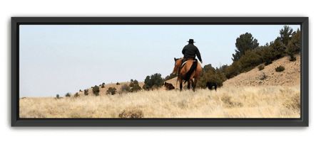 Cowboy on horseback riding through dry grass hills with scattered trees under a clear sky, set in a black wood frame