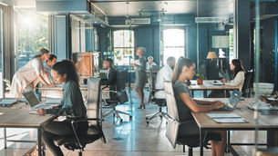 People working at desks in a modern, glass-walled office with laptops and papers. Sunlight streams in, creating a focused, busy atmosphere.