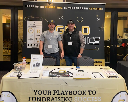 Brian Ludwig and Robby Muniak Fundraising Coaches smile in front of Gold Athletics Fundraising Display at a coaches conference