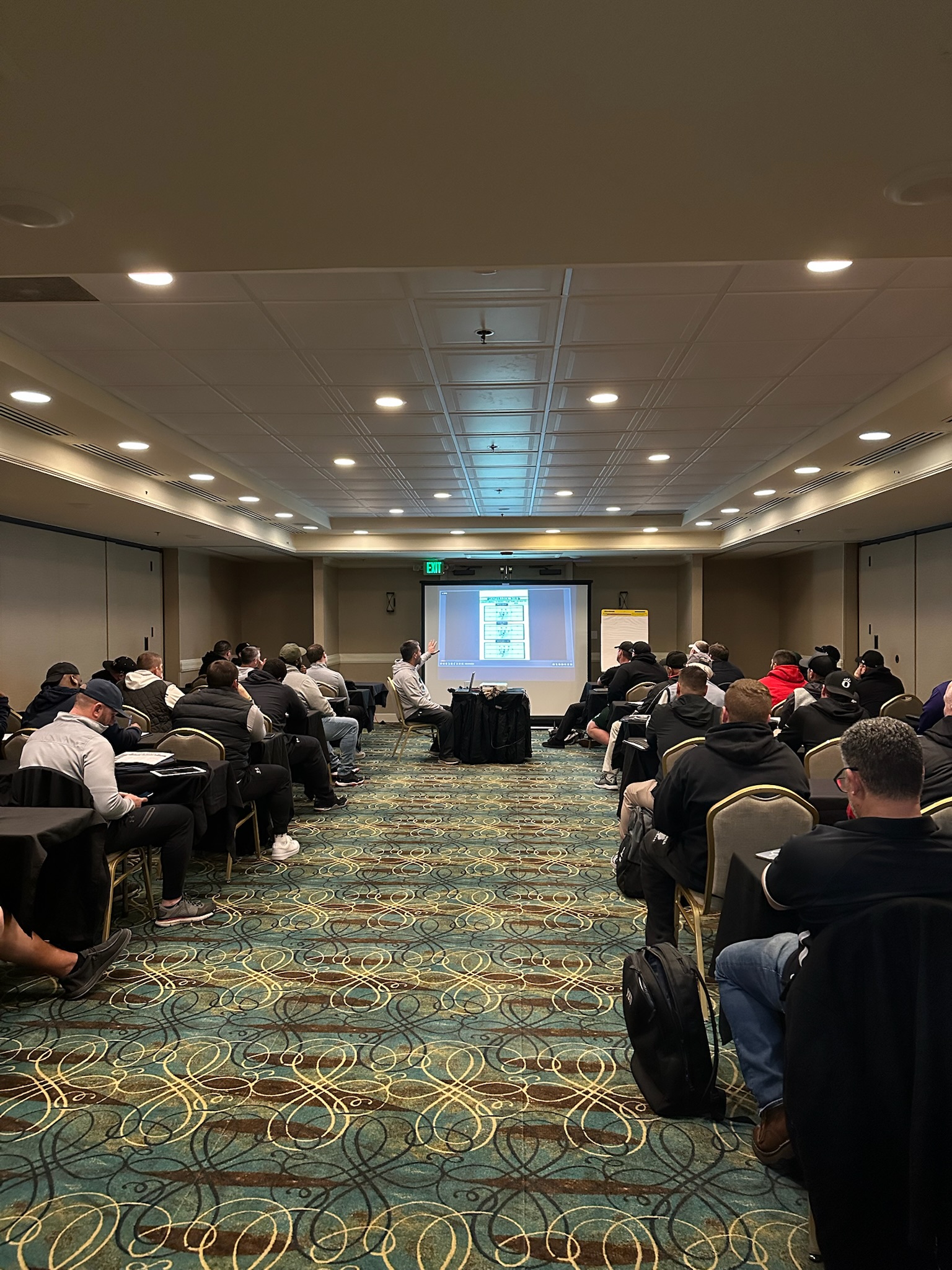 People in a conference room watch a presentation on a projector screen. Chairs are arranged in rows on a patterned carpet. Sports Fundraising, coaching clinic, Gold Athletics