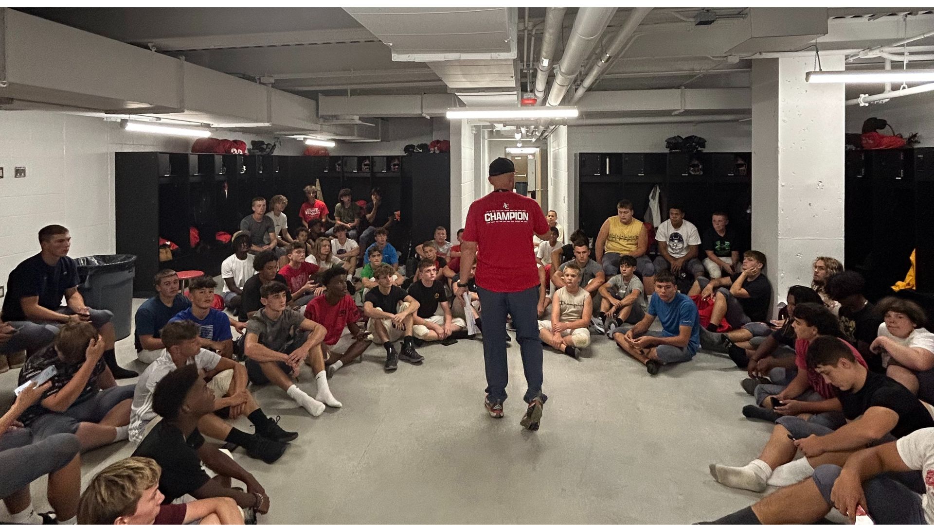 A group of people sit on a locker room floor, attentively listening to a person in a red shirt labeled 
