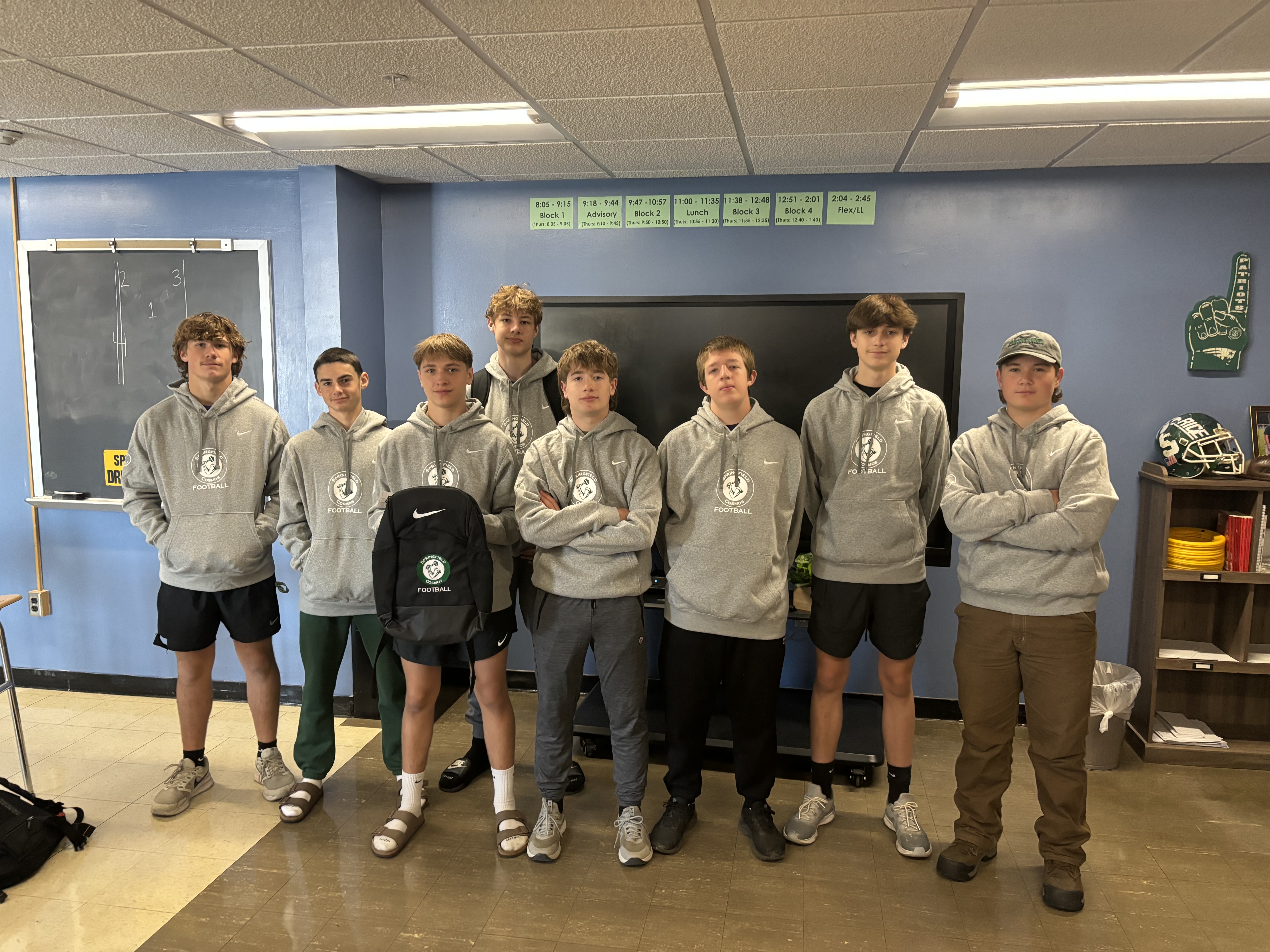 Eight young men in matching gray hoodies pose in a classroom. A blackboard, sign, and football helmet are visible. They appear relaxed. Gold Athletics Fundraising, Fundraising Prizes