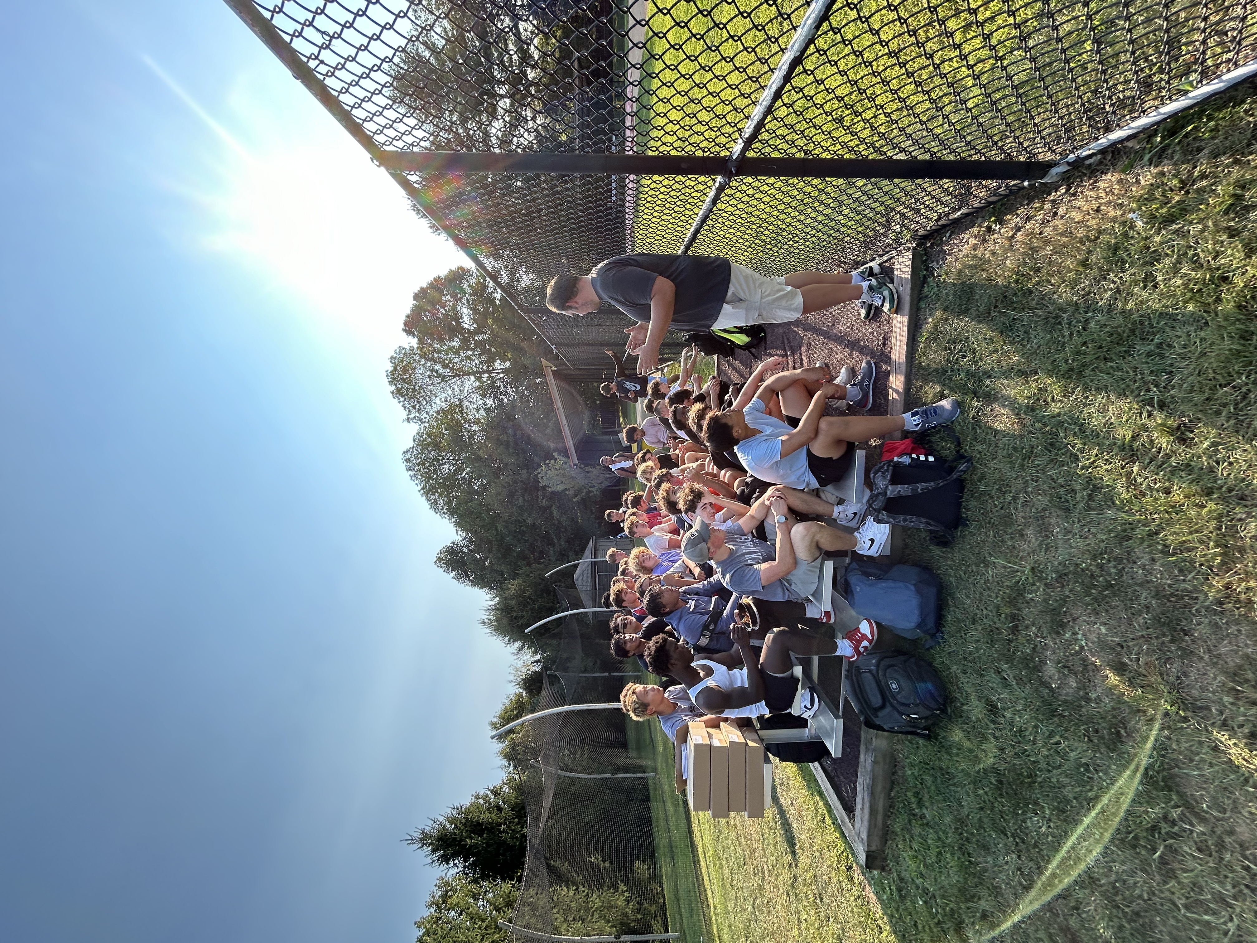 A group of people sit on bleachers outdoors, listening to a standing speaker. Sunny day, chain-link fence, and trees in the background. Gold Athletics, fundraising