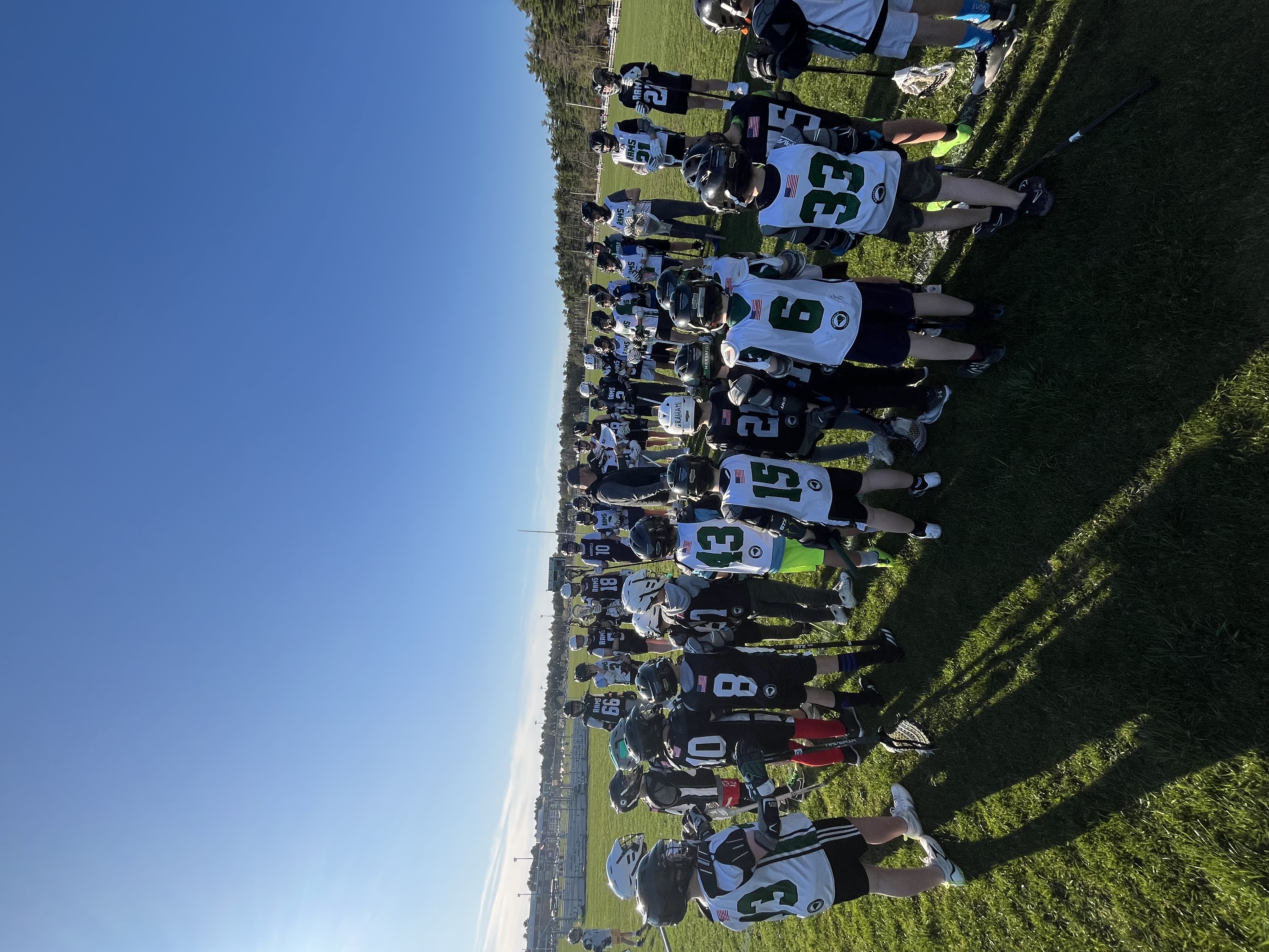 Children in lacrosse gear gather on a grassy field, listening intently to a coach. Bright jerseys with numbers stand out against clear blue sky. Gold Athletics, fundraising.