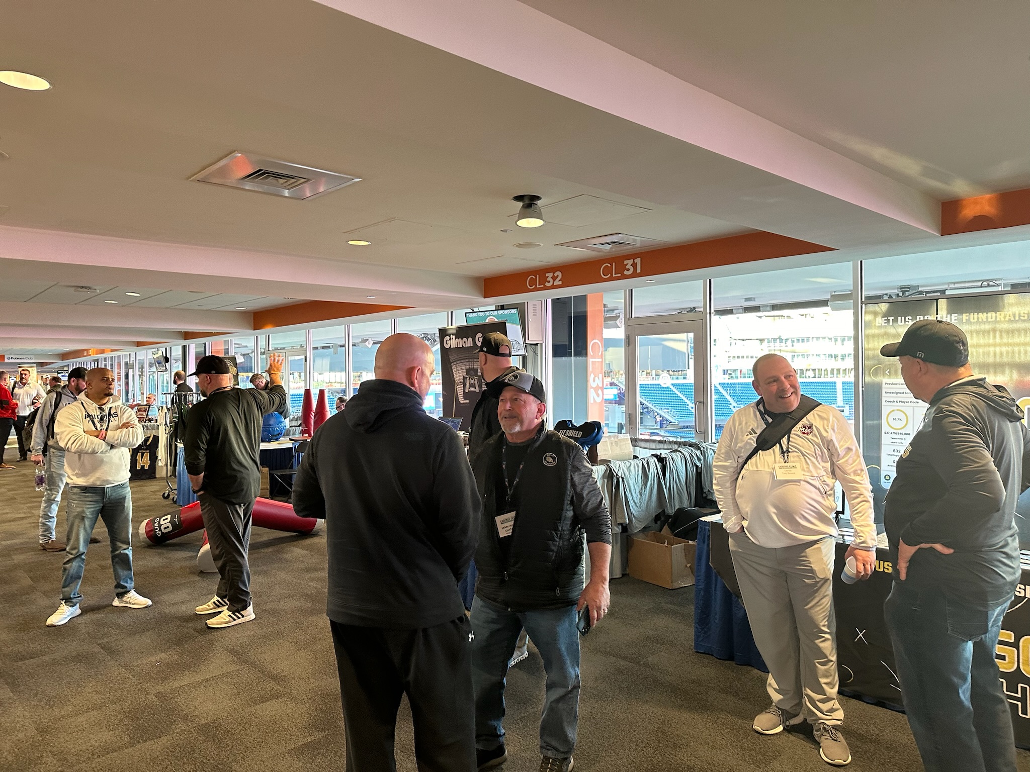 People converse in a carpeted indoor space with a window view of a stadium. Casual, friendly atmosphere. Gold Athletics, Coaches Clinic, Sports fundraising, coaches and representatives collaborating