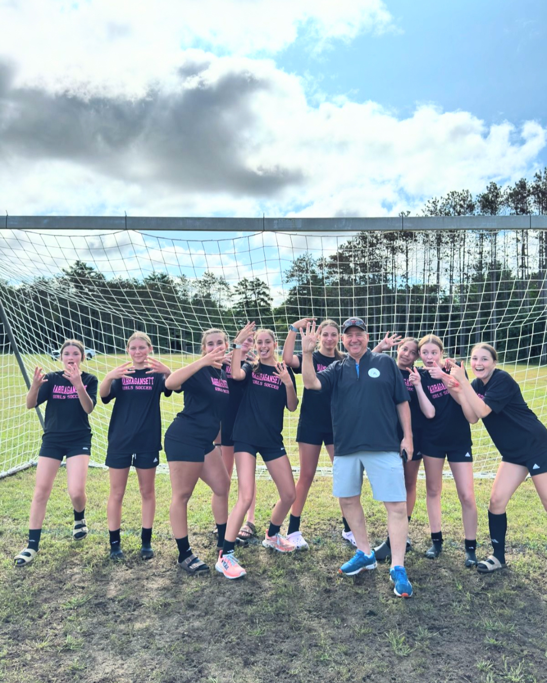 Soccer team of eight in black shirts and shorts, posing and smiling by a goalpost on a field. Coach in blue shoes joins. Overcast sky. Gold Athletics, fundraising, Steve Ash