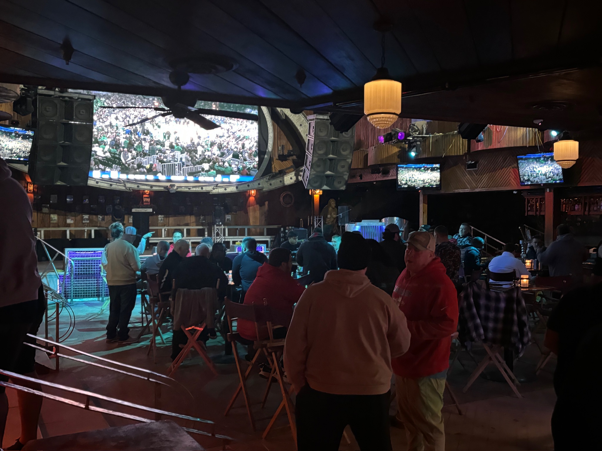 People gather in a dimly lit bar, watching a sports event on multiple screens. Warm lighting and casual attire create a relaxed atmosphere. Sports Fundraising, coaches clinic, Gold Athletics