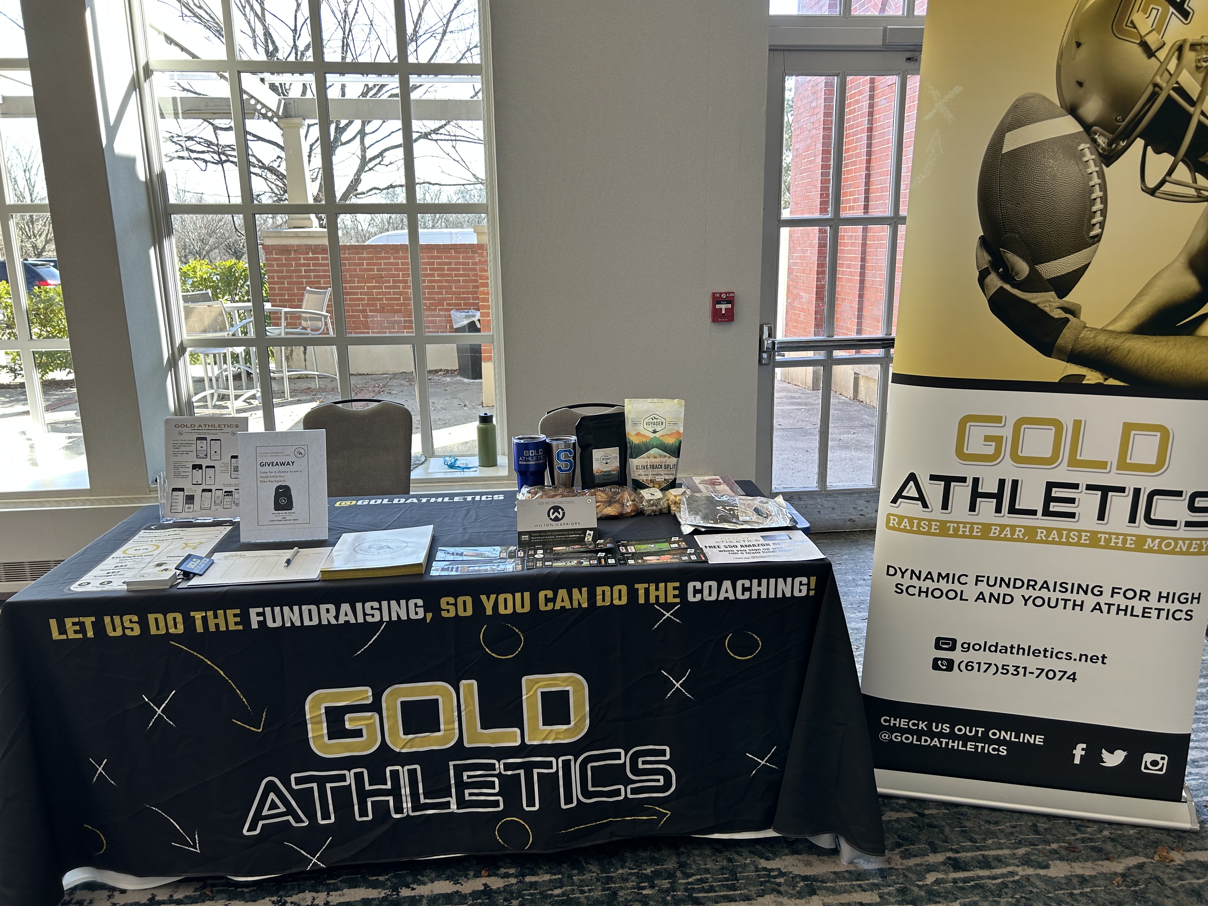 Gold Athletics booth with promotional materials, a large banner, and a giveaway sign. Sunlit window in the background. Sports Fundraising collaboration event