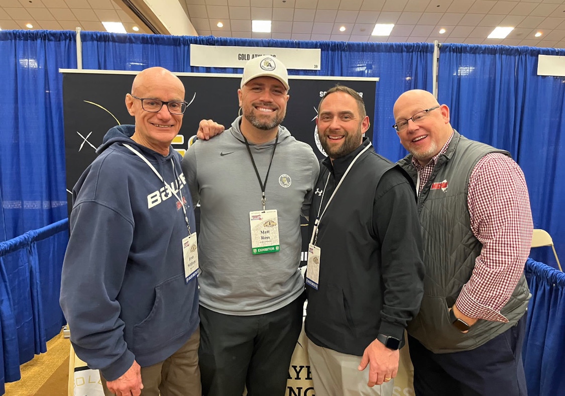 Four men smiling at a booth with blue curtains in the background. They wear name tags and casual attire, exuding a cheerful mood. Sports Fundraising, MSSADA, Gold Athletics