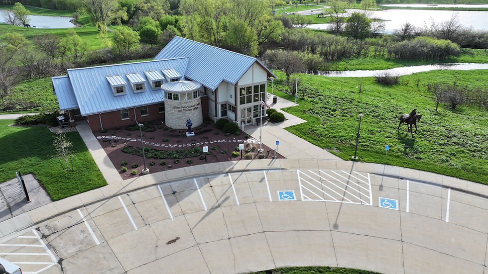Drone image of a visitor building with a parking lot and  panther statue and a public art piece in front of the building and green prairie land, green trees and water in the background.